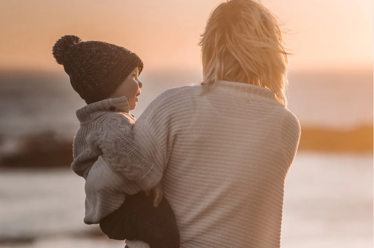 Parent holding a young child in a knit hat, looking out at sunset